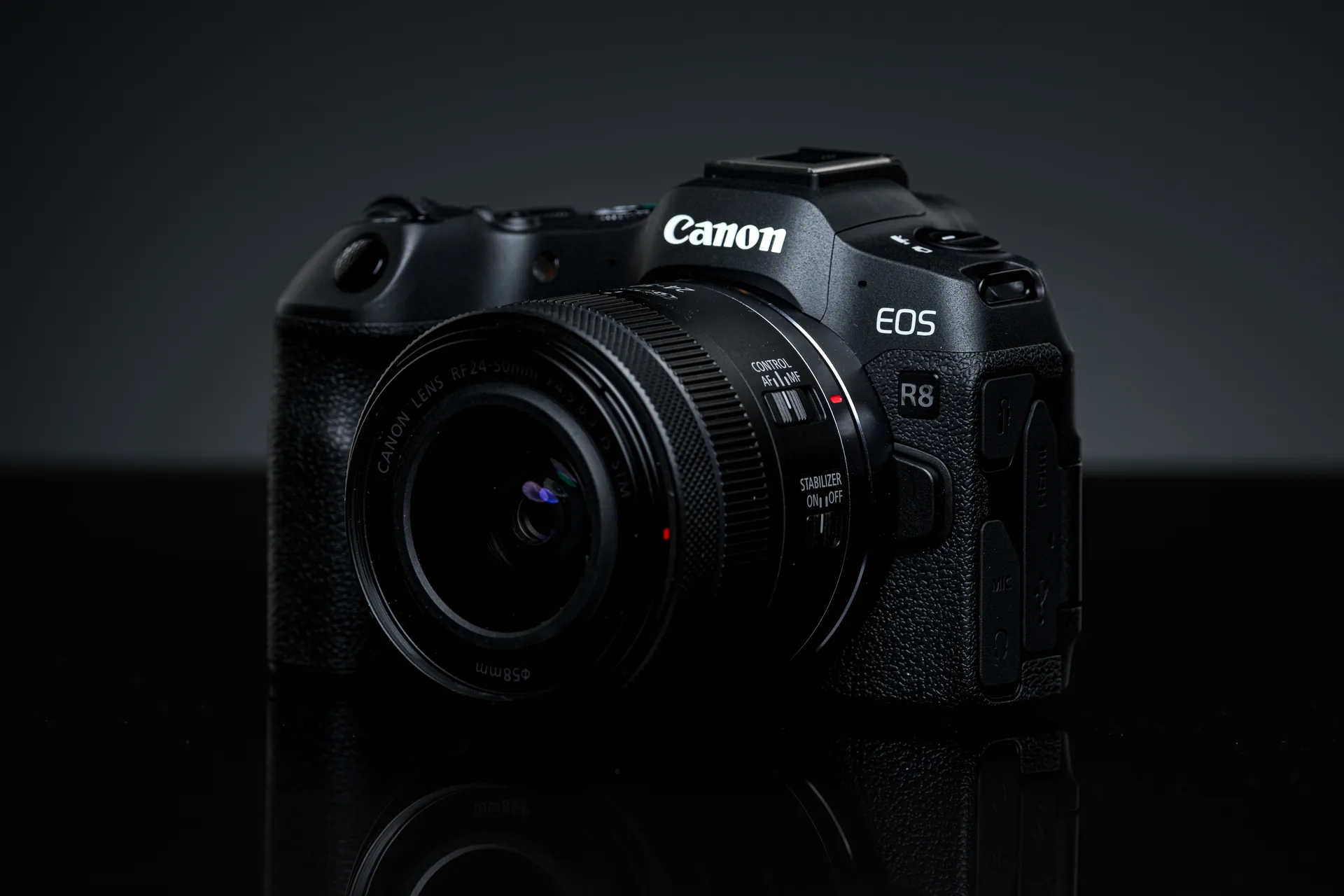 A black Canon EOS R8 with an RF zoom lens, on a dark reflective surface