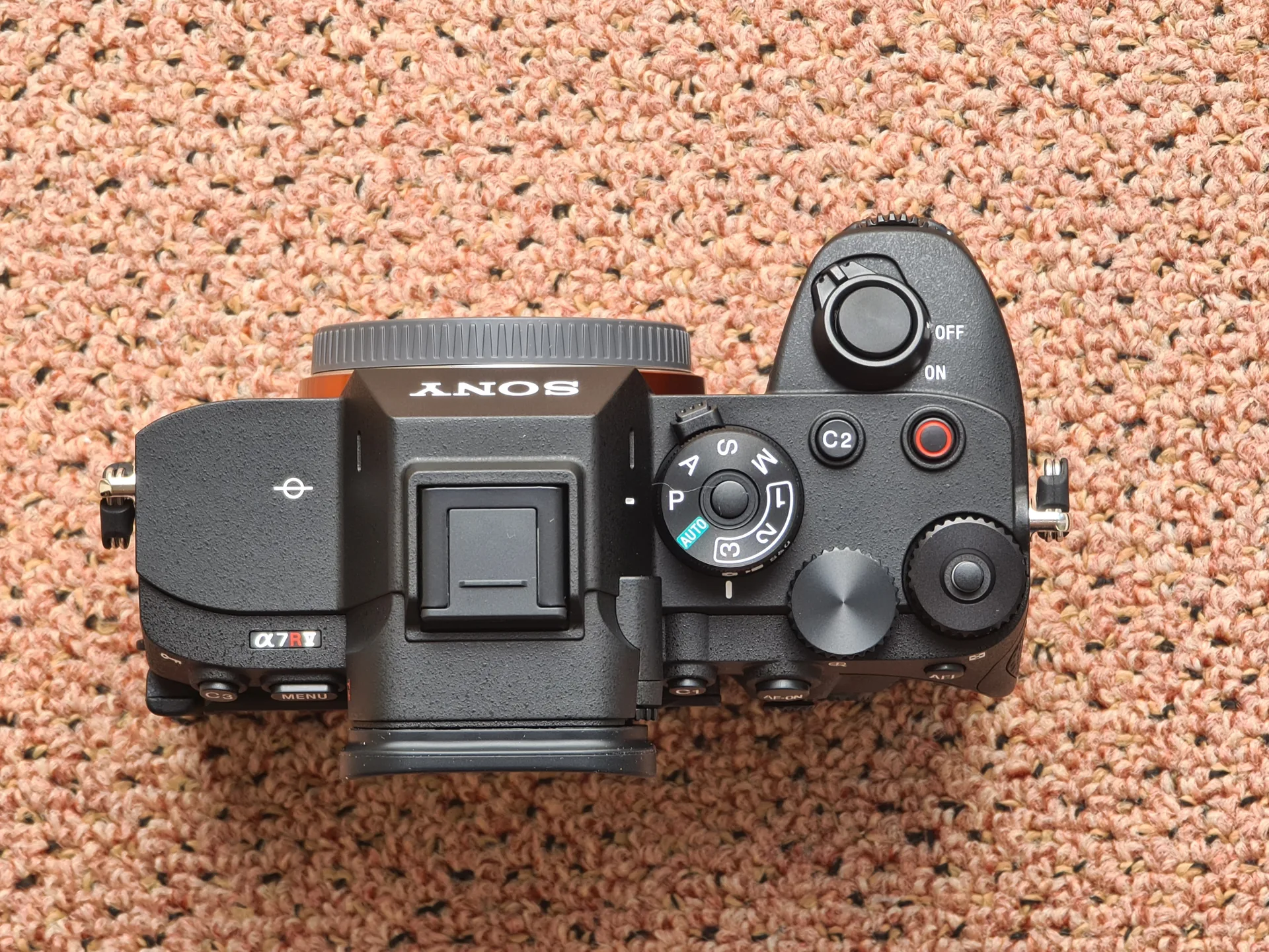 A black Sony a7R V body seen from directly above on a textured orange surface, showing its dials and buttons