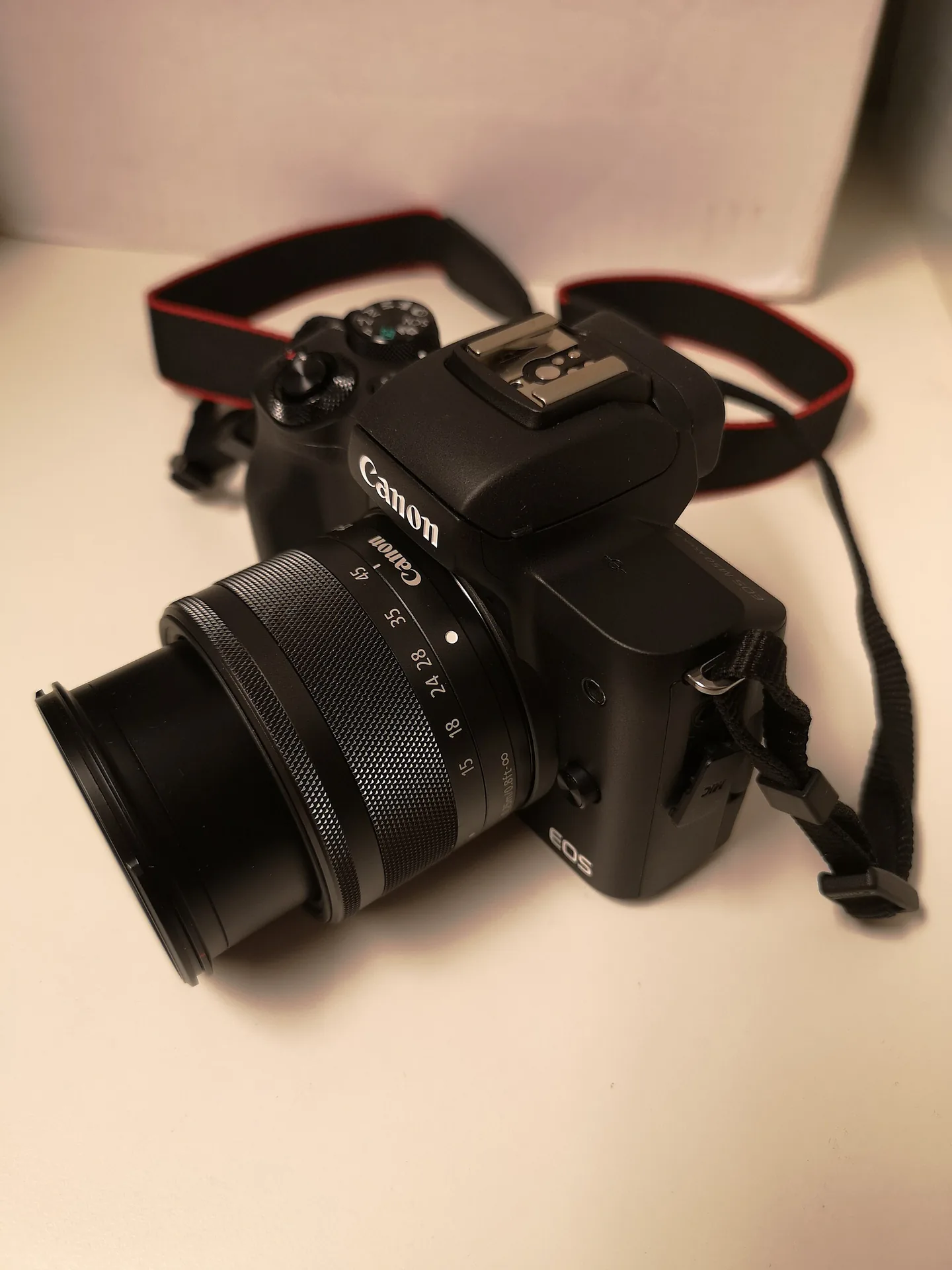 A black Canon EOS M50 Mark II with a zoom lens and its strap, standing on a table against a pale wall