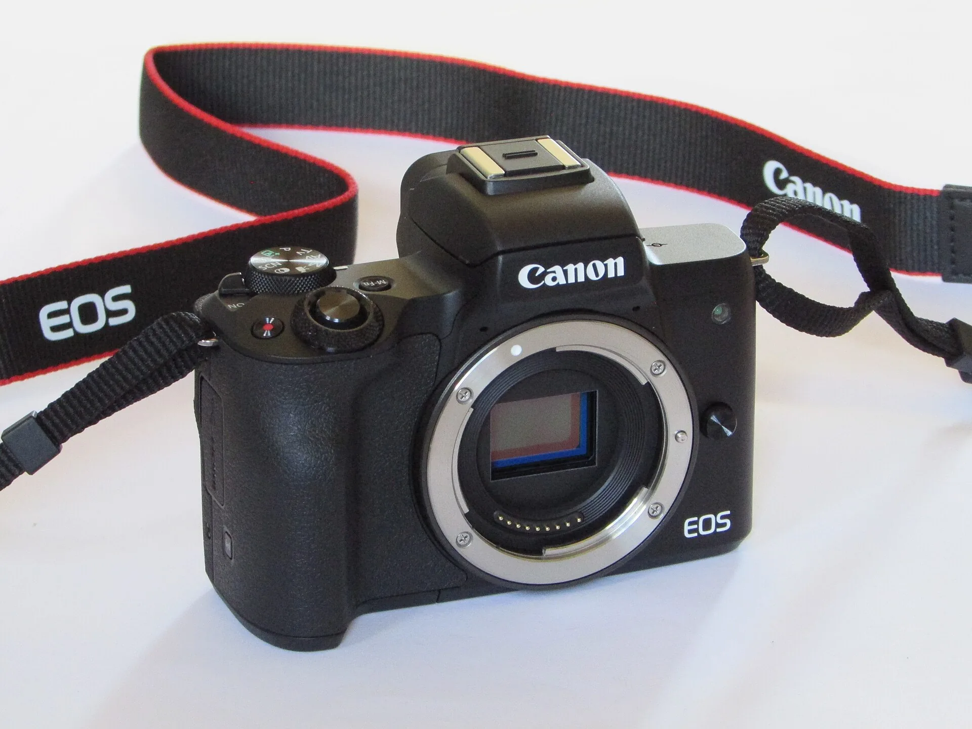 A black Canon EOS M50 body with no lens fitted and a red-trimmed Canon strap, seen from the front on a white background