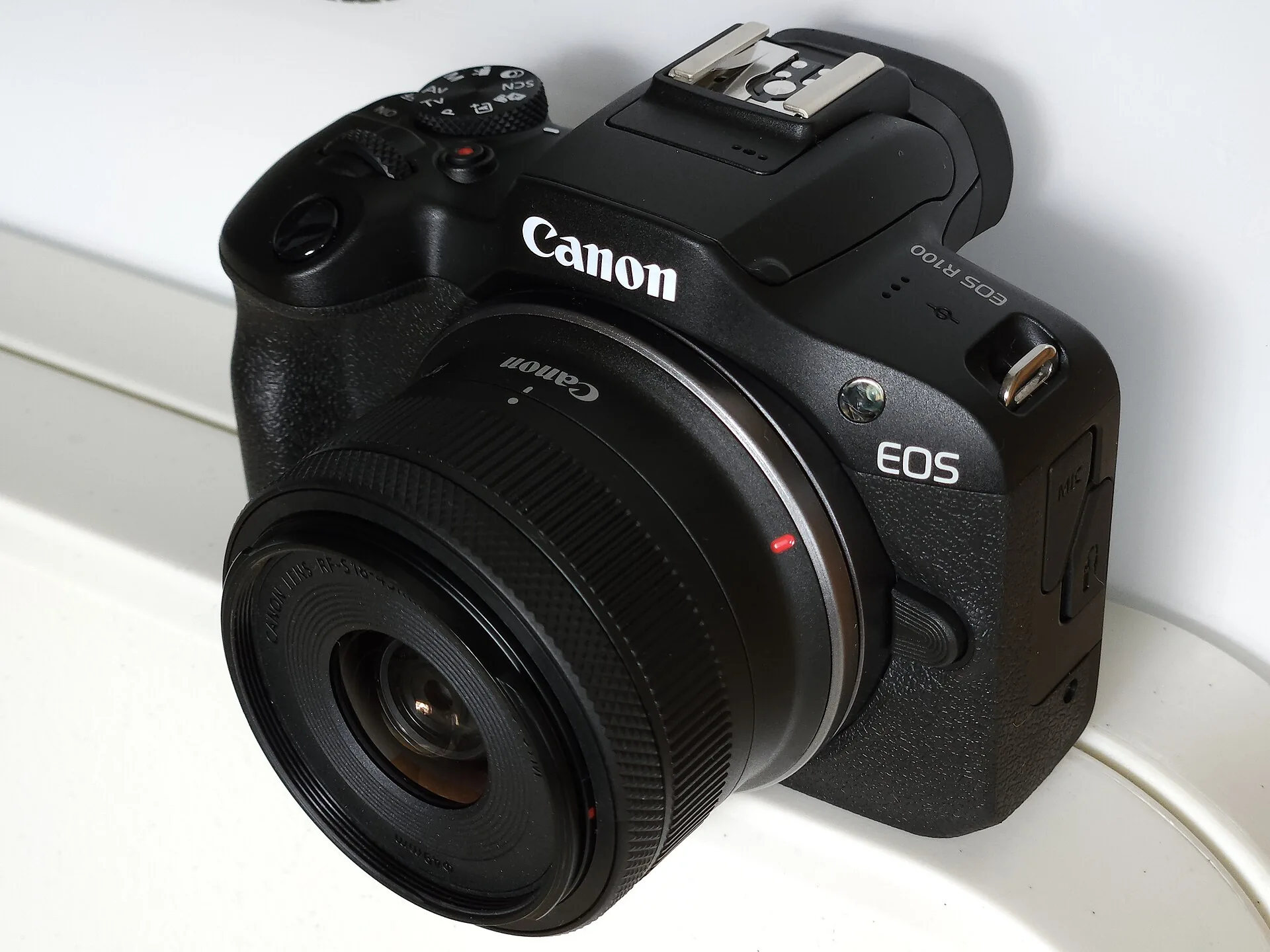 A black Canon EOS R100 with the RF-S 18-45mm kit zoom lens, seen from the front left on a white surface